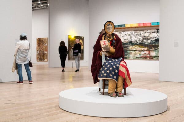 A multimedia sculpture portrays a Madonna figure with a blanket as a shawl and flag wrapped around her knees.