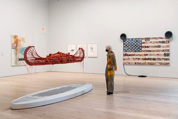 A visitor inspects a red canoe that's made of wood and filled with environmental detritus. Back right, Quick-to-See Smith's